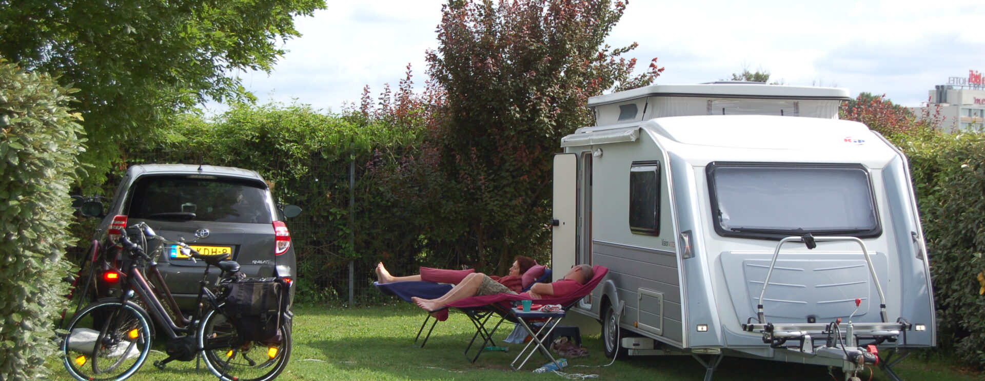 Un couple fait la sieste dans des fauteuil relax devant leur caravane stationnée avec leur voiture sur un emplacement d'herbe du camping La Durance de Cavaillon. Leurs vélos électriques sont visibles sur la photo