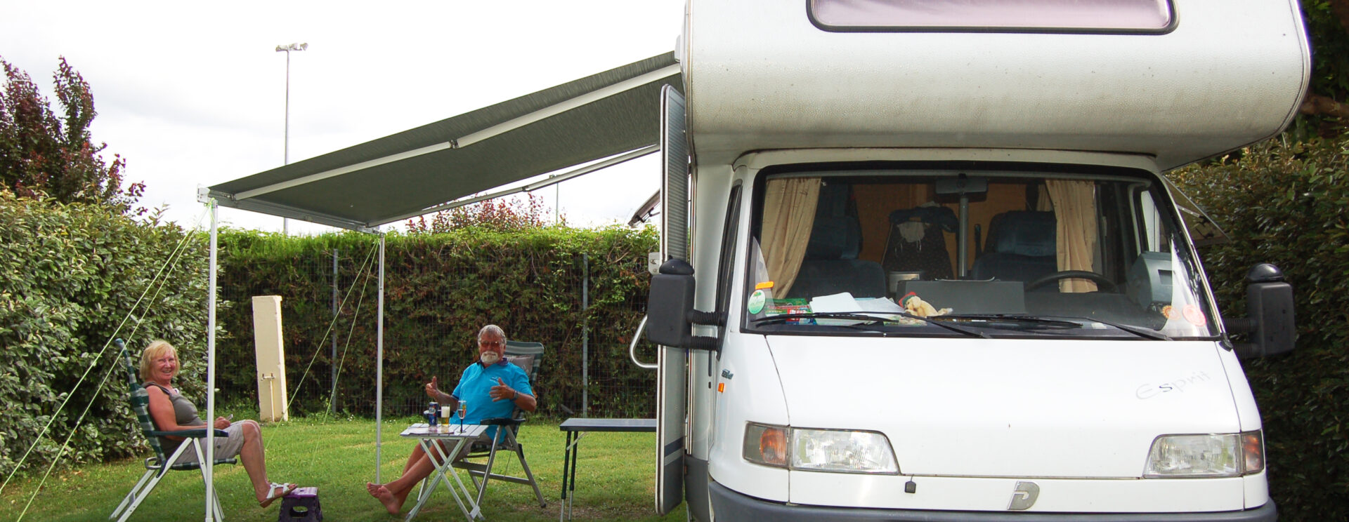 Deux campeurs (un couple) prennent l'apéritif sur une table pliante, assis dans des fauteuils relax, devant leur camping-car stationné sur un emplacement herborisé du camping La Durance de Cavaillon